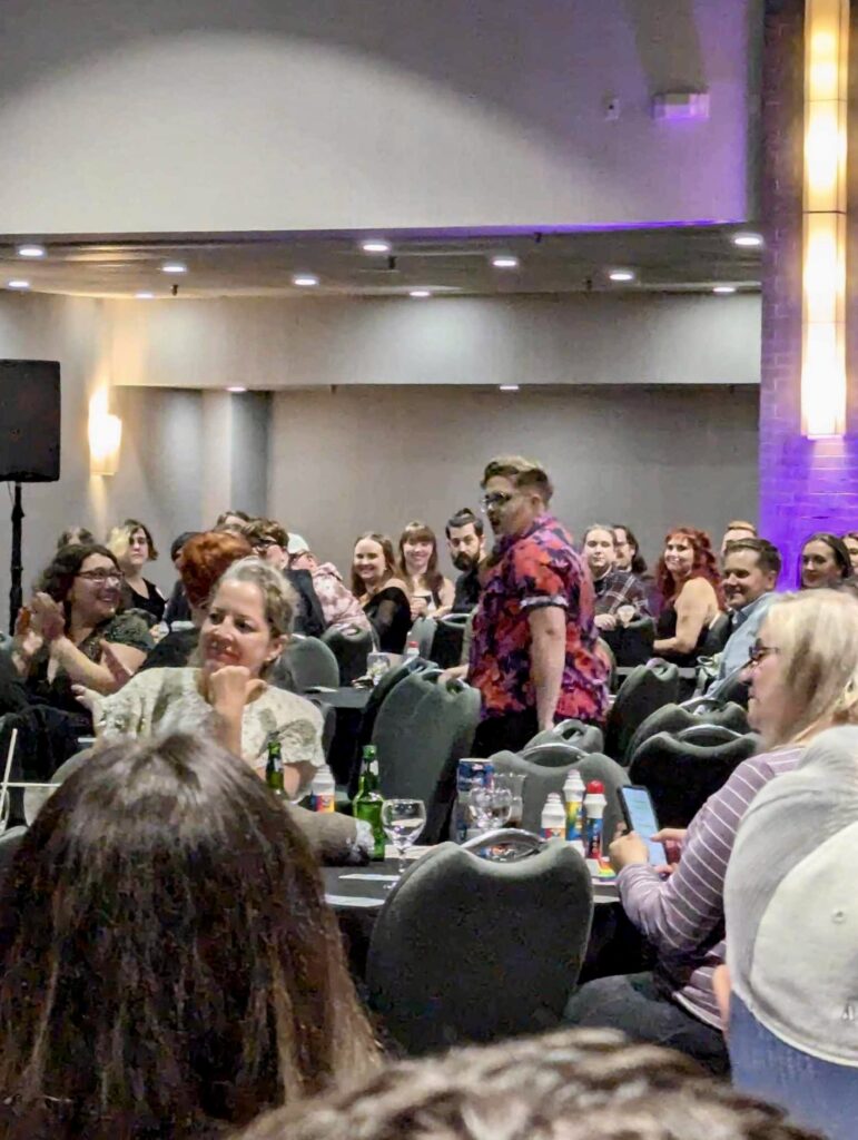 Tori in drag dances in the middle of a crowded room of banquets tables