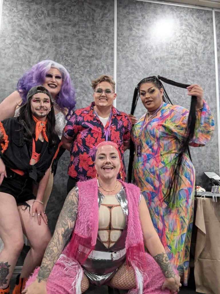 Five drag performers huddle together wearing flamboyant clothes, dramatic wigs and big smiles